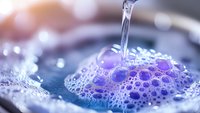 Stream of clear water pouring into purple soap bubbles and foam creating delicate macro texture with bokeh effect and soft lighting for bathroom and spa concepts.