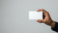 Close-up of male hand with dark skin tone holding blank white business card or paper against neutral gray background for branding and corporate identity mockup.