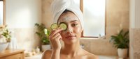 Young woman with towel on head applying cucumber slice to eye for natural skincare routine in bright bathroom with plants.