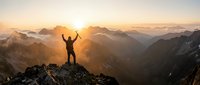 Triumphant hiker with raised arms celebrating success on mountain peak at golden sunrise with dramatic alpine landscape and layered mountain ranges.
