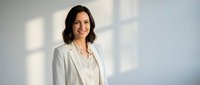 Professional businesswoman in white blazer smiling confidently in modern office with natural lighting for corporate presentations and marketing materials.