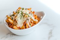 Fresh penne pasta in rich tomato sauce topped with shaved parmesan cheese and herbs, served in white ceramic bowl on marble surface.