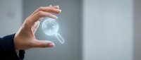 Professional hand holding glowing magnifying glass icon in modern office environment for business analysis and search concepts.