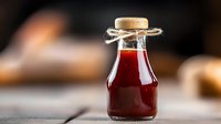 Small glass bottle with red sauce or syrup sealed with cork and twine on wooden surface against blurred rustic background.
