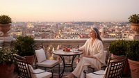 Woman in white robe enjoying peaceful morning coffee on luxury rooftop terrace overlooking historic city skyline at golden hour sunrise for wellness lifestyle.