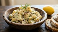 Potato salad with fresh dill and creamy dressing served in rustic wooden bowl, accompanied by lemon and bread slices for lunch or dinner.
