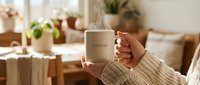 Cozy morning at home with woman in knit sweater holding steaming coffee mug in warm living room with plants and natural light.