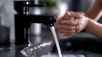Hands washing under running water with splashing droplets in bathroom sink, close-up view showing personal hygiene and cleanliness in dramatic lighting.