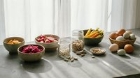Healthy organic ingredients arranged on kitchen counter including nuts, seeds, fresh vegetables and fruits for nutritious meal preparation and cooking.