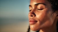 Young woman with freckled skin and closed eyes enjoying sunlight at golden hour, side profile portrait showing natural beauty and serenity against blurred beach background.