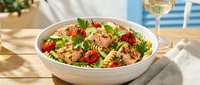 Fresh pasta salad with cherry tomatoes, herbs and vegetables served in white bowl on wooden table with wine glass for summer dining.