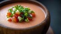 Creamy pink gazpacho soup garnished with fresh diced vegetables and herbs in wooden bowl, close-up view with selective focus on colorful toppings.