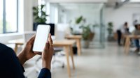 Close-up of hands holding smartphone with blank white screen in modern office workspace with blurred colleagues and plants in background.