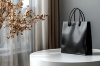 Black leather tote bag on white round table with dried flowers and curtains in background. Luxury fashion accessory for retail, e-commerce, and lifestyle design projects.