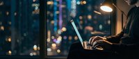 Professional working late at night on laptop with city lights bokeh background creating atmospheric workspace ambiance for productivity and focus concepts.