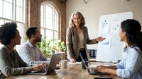 Diverse team of professionals gathered around wooden table during business meeting, with middle-aged woman presenting strategy on whiteboard near large windows and potted plant in modern office.