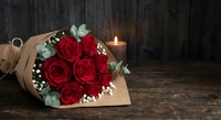 Red roses bouquet wrapped in kraft paper with eucalyptus and baby's breath on rustic wooden background with candle light for romantic occasions.
