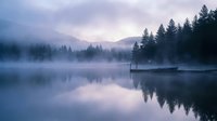 Misty lake at dawn with wooden dock and pine forest silhouettes reflected in calm water. Serene mountain landscape with fog rolling over trees creates peaceful atmosphere.