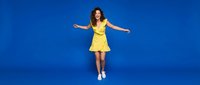 Young Caucasian woman in bright yellow dress dancing with arms outstretched against vibrant blue studio background for lifestyle and fashion content.