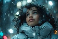 Young mixed race woman with curly hair looking up at falling snow, wearing winter jacket against blurred bokeh lights and moody teal atmosphere.