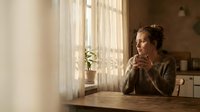 Young Caucasian woman in cozy sweater sitting at wooden table by window with warm sunlight, holding coffee mug in peaceful morning kitchen interior.
