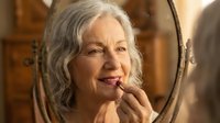 Senior Caucasian woman applying lipstick while looking in vintage mirror, showcasing mature beauty and self-care routine in warm indoor setting.
