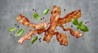 Crispy bacon strips with fresh basil leaves and black peppercorns scattered on gray concrete surface for culinary design and food advertising projects.