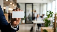Businessman in dark suit holding blank white business card in modern office lobby with colleagues and green plants in background.