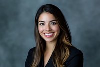 Young Hispanic businesswoman with long ombre hair wearing black blazer smiling confidently against gray studio background, professional corporate headshot portrait.