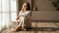 Young woman in white pajamas sitting on wooden floor enjoying morning sunlight streaming through window in cozy bedroom interior for wellness lifestyle.