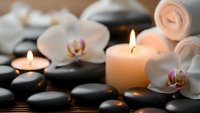 Spa wellness scene with burning candles, white orchid flowers, black hot stones and rolled towels creating peaceful relaxation atmosphere for massage therapy.