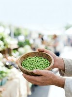 Farmer holding wooden bowl filled with fresh green peas at outdoor market, displaying harvest produce for sale to customers in blurred background.