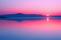 Tranquil lake landscape at sunset with mountain silhouettes against pink and purple sky, mirror reflection on calm water surface creates symmetrical nature view.