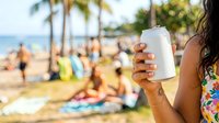 Closeup of woman's hand holding blank white cup at tropical beach with palm trees and ocean in background, sunscreen product mockup for summer travel and vacation marketing.