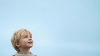 Young boy with blonde curly hair looking upward against clear blue sky, expressing wonder and optimism for aspirational and motivational content.