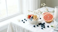 Healthy breakfast bowl with granola, blueberries, strawberries, and grapefruit on a white linen table by a sunny window with spring blossoms.