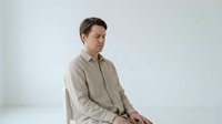 Young Caucasian man in beige linen shirt sitting thoughtfully against white background, minimalist portrait for lifestyle and fashion content.