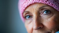 Close up portrait of mature woman wearing pink knitted hat, showing strength and resilience during cancer treatment and recovery journey. Soft natural lighting highlights blue eyes.