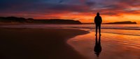 Silhouetted male figure standing on wet sand beach at dramatic sunset with golden and orange sky reflected in water, contemplative mood.