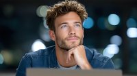 Young handsome Caucasian man with curly hair and beard smiling thoughtfully with hand on chin against blurred city lights background at night.