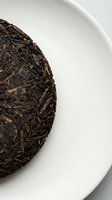 Compressed dark tea cake with aged leaves on white ceramic plate, isolated against neutral background for beverage and wellness design projects.