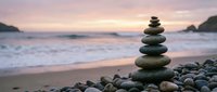 Balanced stone cairn on pebble beach at sunset with soft waves and mountains creating peaceful meditation scene for wellness and mindfulness concepts.