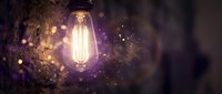 Vintage Edison bulb glowing with warm filament against dark background with magical bokeh lights and sparkling particles creating atmospheric mood.