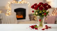Red roses in crystal vase on white table with fireplace and warm lights creating romantic atmosphere for Valentine's Day or anniversary celebration.