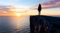 Woman silhouette standing on dramatic cliff edge overlooking ocean at golden sunset. Adventure travel and freedom concept for inspirational content.