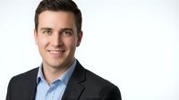 Professional young Caucasian businessman in dark suit and blue shirt smiling confidently at camera against white background for corporate headshots.