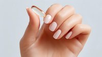 Female hand with elegant beige manicure holding transparent glass dropper against light background, demonstrating skincare or beauty product application.