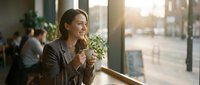 Young professional woman enjoying morning coffee by cafe window with warm sunlight, relaxed business casual lifestyle moment for workplace wellness concepts.