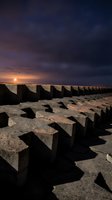 Concrete breakwater blocks arranged in rows along sandy beach at sunset with dramatic cloudy sky and golden light reflecting on water.