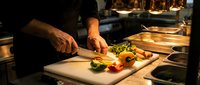 Chef chopping fresh colorful vegetables on a white cutting board in a professional restaurant kitchen with warm dramatic lighting.